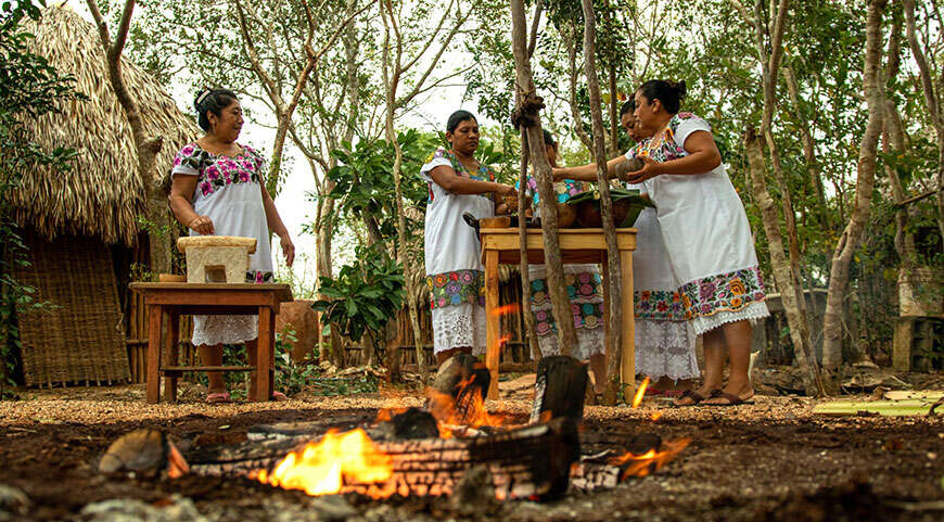 Four women in traditional embroidered dresses attend an outdoor cooking class in Mexico, with a campfire in the foreground.