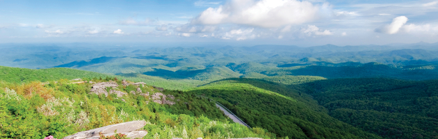 A vast view of the lush, green Southern Appalachian Mountains in Tennessee seen from a rocky overlook under a blue, cloudy sky.