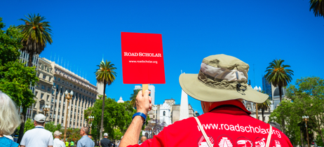 A leader in a red Road Scholar t-shirt holding a red Road Scholar sign while walking through a city