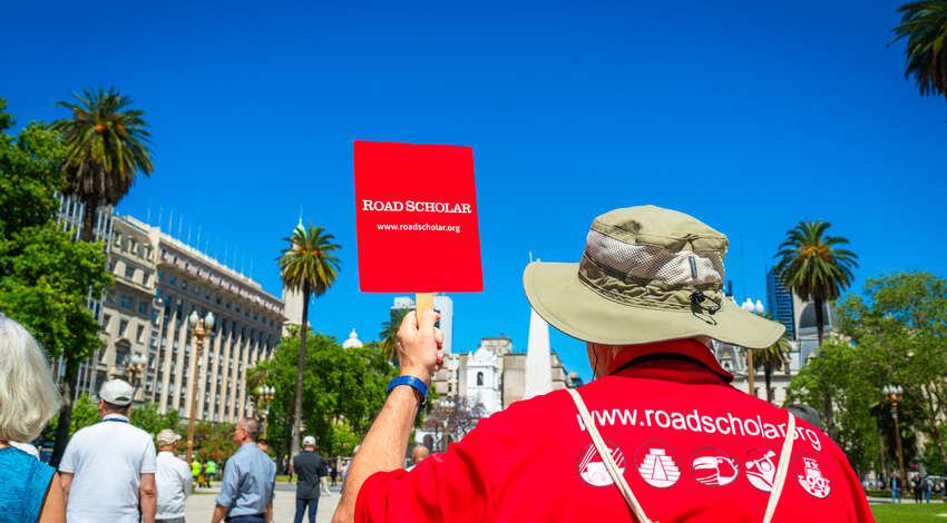 A leader in a red Road Scholar t-shirt holding a red Road Scholar sign while walking through a city