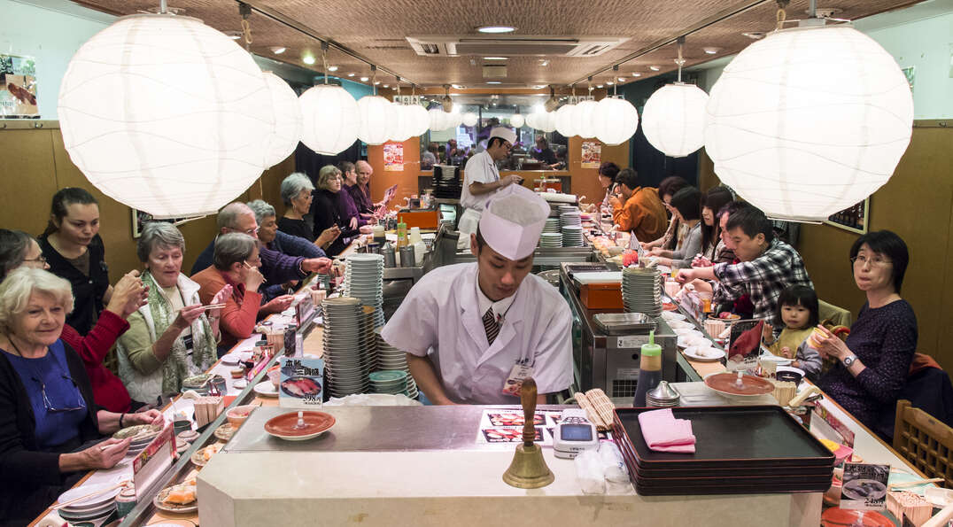 Diners sit along a conveyor belt at a sushi restaurant in Japan as chefs prepare food under large, spherical white paper lanterns.