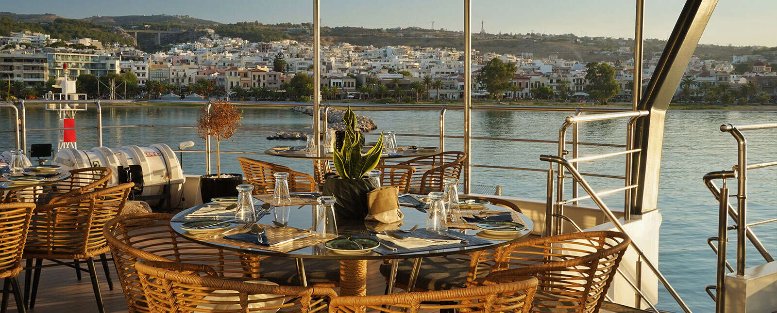 Outdoor dining area on a boat deck with set tables and wicker chairs overlooking a coastal town at sunset.