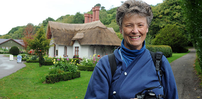 A smiling woman holding a camera stands on a path in front of a traditional thatched-roof cottage and garden in Ireland.