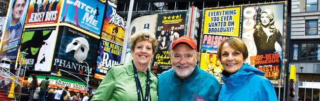 Three people smiling in Times Square, New York, with vibrant Broadway theater billboards in the background.