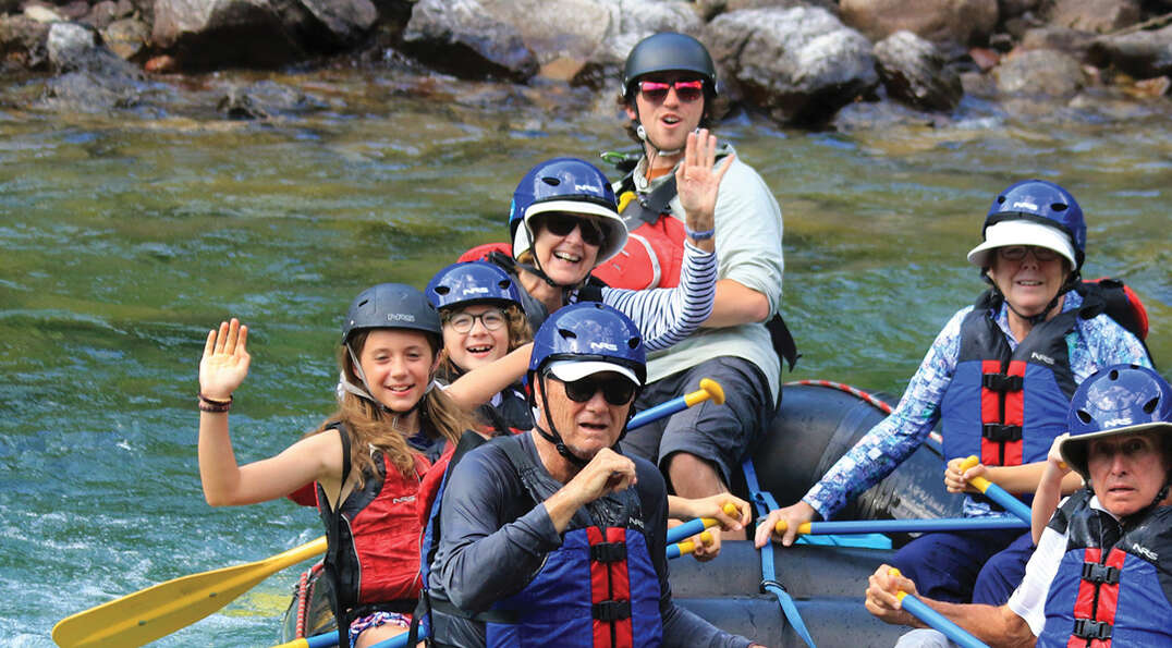 A multi-generational family in helmets and life vests smiles and waves while rafting down a river in Glacier National Park, Montana.