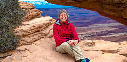 A solo traveler in a red jacket smiles while sitting on a rock formation, with a vast canyon visible in the background.