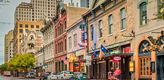 A daytime view of the historic buildings and storefronts lining a busy Sixth Street in Austin, Texas, with cars and pedestrians.
