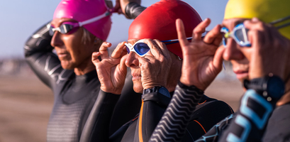 Three people wearing wetsuits and colorful swim caps age adventurously as they adjust their goggles on a beach.