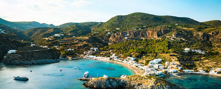 An aerial view of a coastal town in Greece featuring white buildings and boats in a turquoise bay surrounded by rugged green hills.