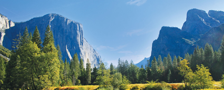 A panoramic landscape of Yosemite Valley featuring massive granite rock formations and evergreen trees under a clear blue sky.