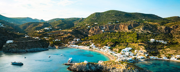 An aerial view of a coastal town in Greece featuring white buildings and boats in a turquoise bay surrounded by rugged green hills.
