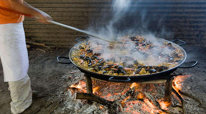 A person prepares traditional Spanish paella, stirring seafood in a large pan as it steams over an open fire.