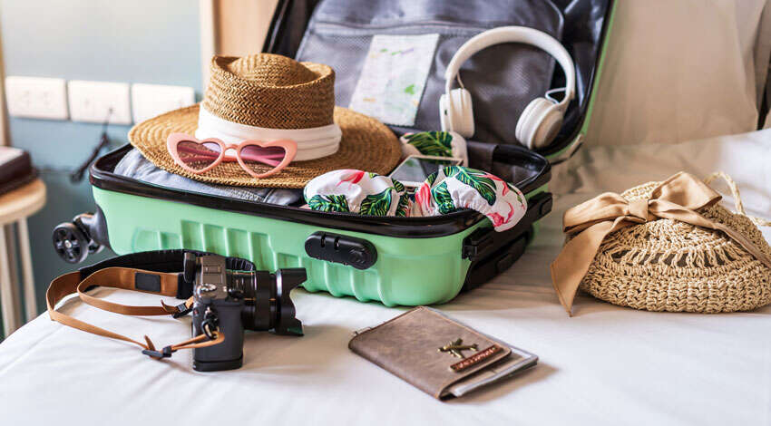 An open mint green suitcase packed for a vacation with a straw hat, heart-shaped sunglasses, and a camera lying on a white bed.