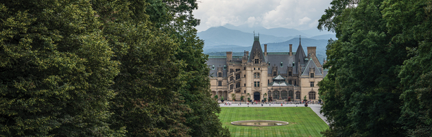 The historic Biltmore Estate in Asheville, North Carolina, framed by lush green trees with a vast lawn and mountain peaks in the distance.