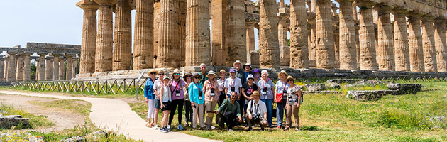 A group of travelers poses for a photo in front of the ancient Greek temples at Paestum in Italy on a sunny day.