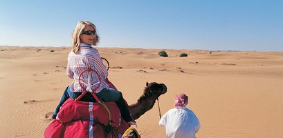 A female solo traveler smiles back at the camera while riding a camel through a vast desert with a guide leading the way.